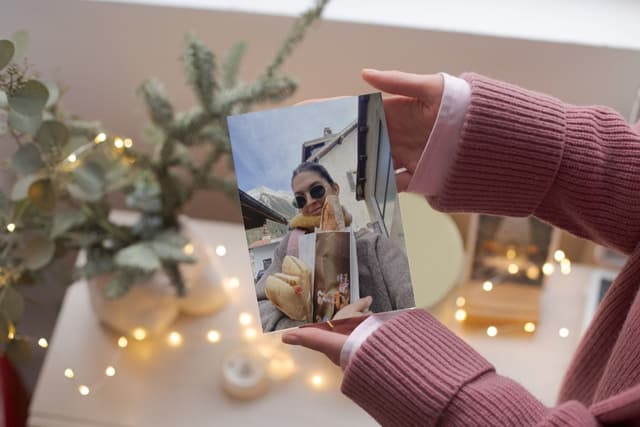 Hands holding the picture of a girl holding a baguette and dressed in wintery style with a background of fairy lights