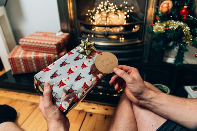 Present wrapped in pastel blue paper with patterns of Santa and reindeers in front of a fireplace with fairy lights in it